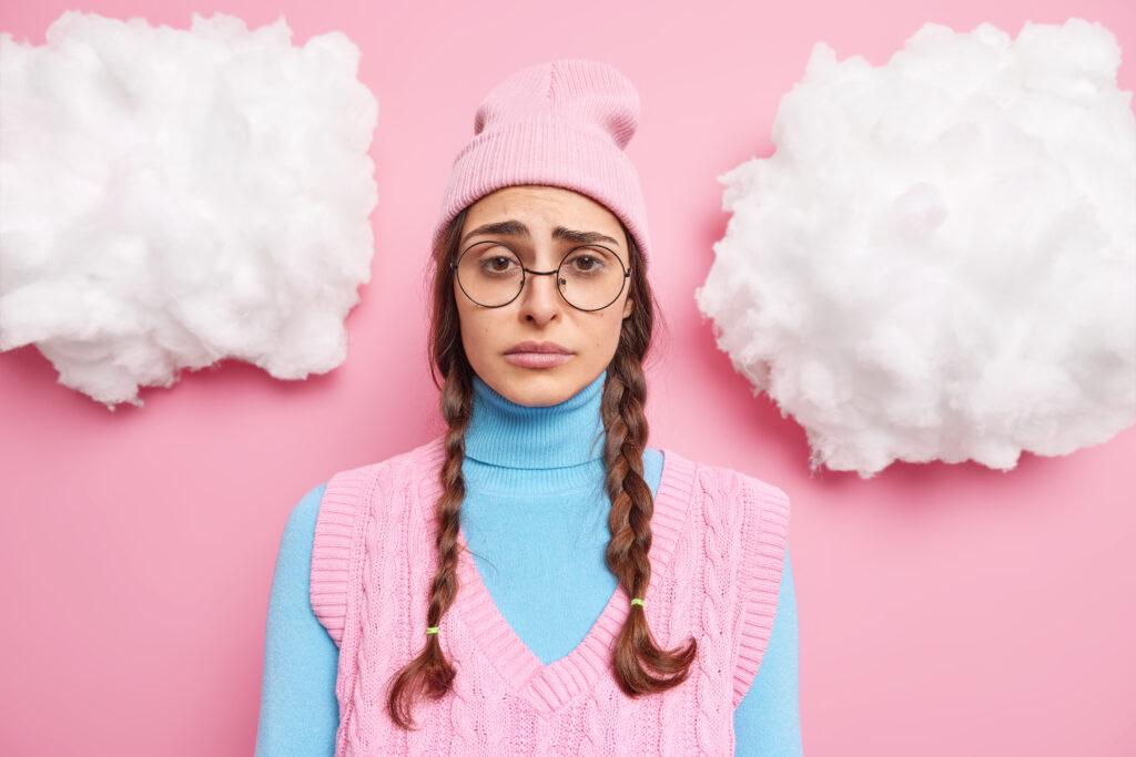 chica con dos coletas tiene expresion sombria enfurrunada y frunciendo el ceno se siente decepcionada o molesta usa gafas redondas sombrero rosa cuello alto y chaleco posa interior 1